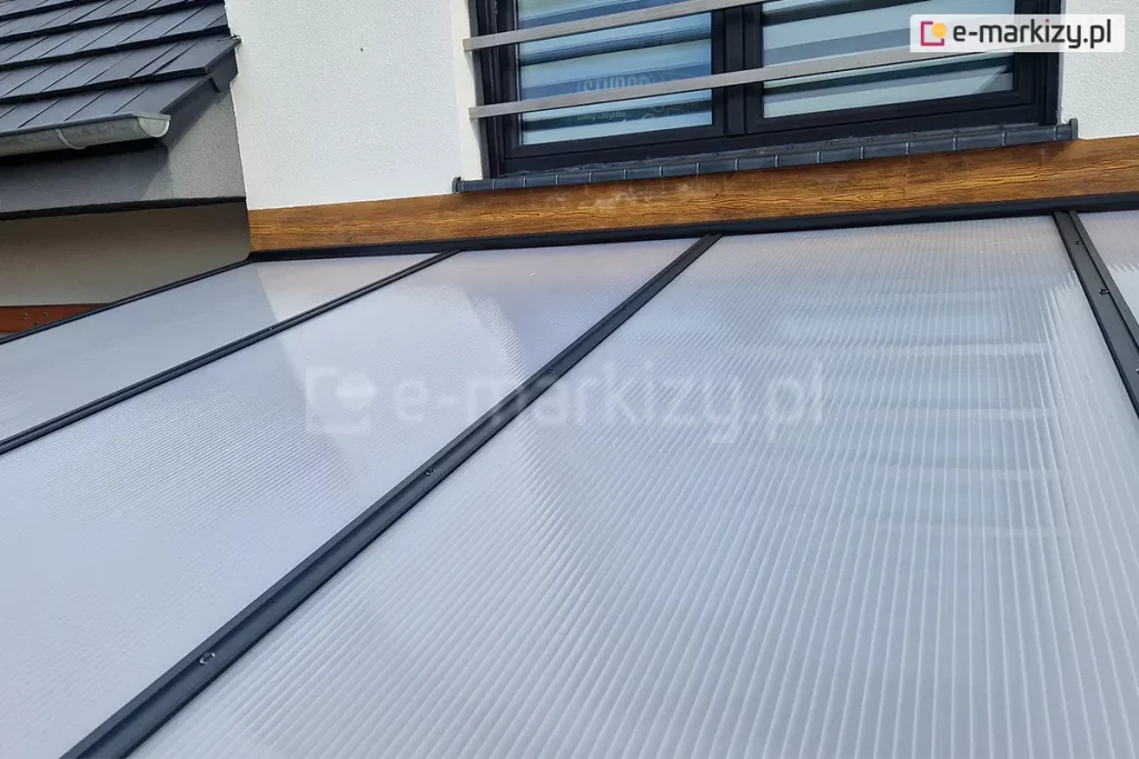 Widok z góry na połać dachu zadaszenia tarasowego Modes w miejscu jego styku z elewacją budynku. Dach tworzą panele z poliwęglanu komorowego o mlecznym zabarwieniu, połączone za pomocą równoległych, antracytowych listew dociskowych z widocznymi punktami mocowań. Górna krawędź konstrukcji jest osadzona w listwie przyściennej, zamontowanej na dekoracyjnym pasie elewacji o strukturze jasnego drewna, tuż pod oknem z barierką francuską. W lewym górnym rogu widoczny jest fragment dachu budynku pokrytego ciemną dachówką oraz system rynnowy.