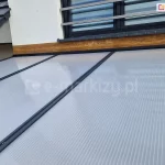 Widok z góry na połać dachu zadaszenia tarasowego Modes w miejscu jego styku z elewacją budynku. Dach tworzą panele z poliwęglanu komorowego o mlecznym zabarwieniu, połączone za pomocą równoległych, antracytowych listew dociskowych z widocznymi punktami mocowań. Górna krawędź konstrukcji jest osadzona w listwie przyściennej, zamontowanej na dekoracyjnym pasie elewacji o strukturze jasnego drewna, tuż pod oknem z barierką francuską. W lewym górnym rogu widoczny jest fragment dachu budynku pokrytego ciemną dachówką oraz system rynnowy.