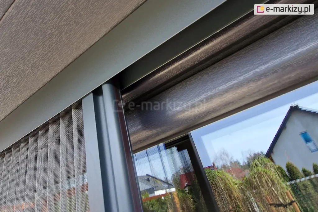 Profil boczny z siatką plisowaną moskitiery Top Zag porusza się wewnątrz górnej prowadnicy zamocowanej do skrzynki rolety zewnętrznej na oknie o antracytowej drewnianej ramie.