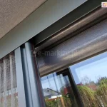 Profil boczny z siatką plisowaną moskitiery Top Zag porusza się wewnątrz górnej prowadnicy zamocowanej do skrzynki rolety zewnętrznej na oknie o antracytowej drewnianej ramie.