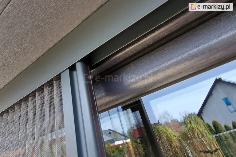 Profil boczny z siatką plisowaną moskitiery Top Zag porusza się wewnątrz górnej prowadnicy zamocowanej do skrzynki rolety zewnętrznej na oknie o antracytowej drewnianej ramie.