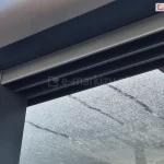 Zbliżenie górnej prowadnicy systemu szyb przesuwnych Sotro zamocowanej do profilu zadaszenia w antracytowym kolorze. W tle widoczne szklane, mocno pokryte skraplającą się parą panele dachowe.