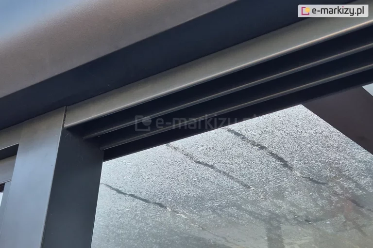 Zbliżenie górnej prowadnicy systemu szyb przesuwnych Sotro zamocowanej do profilu zadaszenia w antracytowym kolorze. W tle widoczne szklane, mocno pokryte skraplającą się parą panele dachowe.