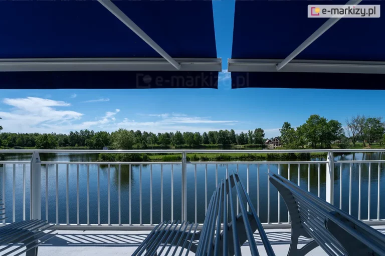 Widok z tarasu lub pokładu jednostki pływającej zabezpieczonego białą barierką o gęsto rozstawionych pionowych szczeblach, za którą rozpościera się panorama jeziora otoczonego gęstą ścianą zielonych drzew pod bezchmurnym, błękitnym niebem. Na pierwszym planie znajdują się srebrne, metalowe ławki o konstrukcji wykonanej z równolegle ułożonych rurek, które ustawione są bokiem do tafli wody. Nad miejscem do siedzenia zamontowane są dwie duże, ciemnoniebieskie markizy tarasowe o gładkim materiale, rozpięte na białych ramionach konstrukcyjnych, które rzucają głęboki cień na strefę wypoczynkową. Tafla wody w jeziorze jest spokojna, odbijają się w niej rosnące na przeciwległym brzegu drzewa oraz pas zielonej roślinności przybrzeżnej. W oddali po prawej stronie widoczne są pojedyncze zabudowania o spadzistych dachach ukryte wśród drzew. Kompozycja zdjęcia dzieli obraz na trzy poziome pasy: ciemny pas markiz u góry, jasny pas krajobrazu z wodą i niebem w środku oraz techniczny pas tarasu z barierką i meblami na dole.