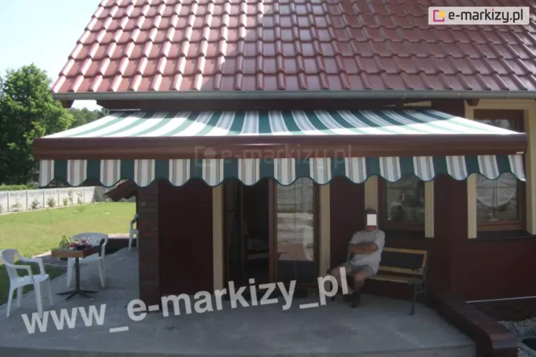 Zdjęcie prezentuje efekt końcowy montażu markizy tarasowej na budynku o ciemnoczerwonej elewacji, pokrytym dachówką ceramiczną w kolorze ceglastym. Markiza jest w pełni rozwinięta, a jej poszycie wykonane jest z materiału w pionowe pasy w kolorach białym, ciemnozielonym oraz szarym. Dolna krawędź tkaniny wykończona jest dekoracyjną, falistą falbaną, która powtarza kolorystykę i wzór głównej połaci. Konstrukcja markizy, w tym belka frontowa oraz kaseta, wykonana jest w kolorze ciemnobrązowym, co współgra z barwą elewacji oraz stolarki okiennej. Pod zadaszeniem, na betonowym tarasie, znajduje się strefa wypoczynkowa z drewnianą ławką, na której siedzi mężczyzna. Po lewej stronie tarasu ustawione są białe, plastikowe krzesła ogrodowe oraz mały stolik. W tle widoczny jest zadbany trawnik, niskie ogrodzenie posesji oraz ściana zielonych drzew pod jasnym niebem. Markiza skutecznie zacienia wejście tarasowe i okna parteru, tworząc komfortową przestrzeń do odpoczynku na zewnątrz.