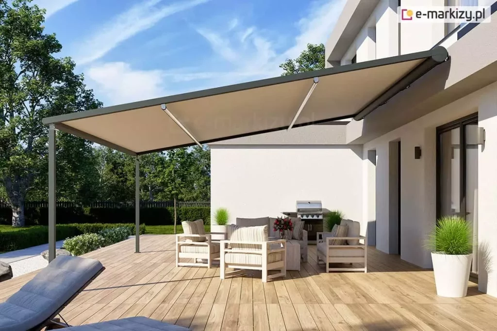 Widok z boku na nowoczesną, lekką pergolę tarasową przyścienną V-Ziiip Selt o konstrukcji aluminiowej w kolorze antracytowym, zamontowaną do jasnej elewacji piętrowego budynku o minimalistycznej bryle. Zadaszenie stanowi gładkie, napięte poszycie tkaninowe w kolorze beżowym, które jest całkowicie rozpięte pod skosem i wsparte na smukłym profilu pionowym przymocowanym do drewnianego podłoża. Pod pergolą, na przestronnym tarasie z jasnobrązowych desek kompozytowych, zaaranżowano strefę relaksu z zestawem mebli ogrodowych w paski oraz profesjonalnym grillem gazowym ze stali nierdzewnej ustawionym pod ścianą. W narożnikach tarasu oraz przy wejściu do domu znajdują się nowoczesne, wysokie donice w kolorze białym z dekoracyjną, zieloną trawą. W tle widoczny jest zadbany ogród z zielonym trawnikiem, niskim żywopłotem oraz drzewami liściastymi pod błękitnym niebem z delikatnymi chmurami. Zdjęcie wykonane w pełnym świetle dziennym, prezentuje estetyczne wykorzystanie lekkich systemów zadaszeń do stworzenia funkcjonalnej jadalni na świeżym powietrzu.