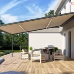 Widok z boku na nowoczesną, lekką pergolę tarasową przyścienną V-Ziiip Selt o konstrukcji aluminiowej w kolorze antracytowym, zamontowaną do jasnej elewacji piętrowego budynku o minimalistycznej bryle. Zadaszenie stanowi gładkie, napięte poszycie tkaninowe w kolorze beżowym, które jest całkowicie rozpięte pod skosem i wsparte na smukłym profilu pionowym przymocowanym do drewnianego podłoża. Pod pergolą, na przestronnym tarasie z jasnobrązowych desek kompozytowych, zaaranżowano strefę relaksu z zestawem mebli ogrodowych w paski oraz profesjonalnym grillem gazowym ze stali nierdzewnej ustawionym pod ścianą. W narożnikach tarasu oraz przy wejściu do domu znajdują się nowoczesne, wysokie donice w kolorze białym z dekoracyjną, zieloną trawą. W tle widoczny jest zadbany ogród z zielonym trawnikiem, niskim żywopłotem oraz drzewami liściastymi pod błękitnym niebem z delikatnymi chmurami. Zdjęcie wykonane w pełnym świetle dziennym, prezentuje estetyczne wykorzystanie lekkich systemów zadaszeń do stworzenia funkcjonalnej jadalni na świeżym powietrzu.