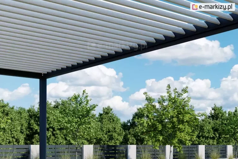 Zbliżenie na techniczne detale zadaszenia nowoczesnej pergoli bioklimatycznej, ukazujące mechanizm ruchomych lameli. Aluminiowe lamele w kolorze białym są ustawione pod skosem w pozycji półotwartej, co demonstruje możliwość precyzyjnej regulacji kąta nachylenia w celu kontrolowania dopływu światła słonecznego oraz wentylacji. Lamele są osadzone w solidnej, poziomej ramie konstrukcyjnej o ciemnoantracytowej barwie, która kontrastuje z ich jasnym kolorem. W lewym dolnym rogu widoczny jest fragment pionowego słupa nośnego, również w kolorze antracytowym, podtrzymującego całą konstrukcję dachu. Tło zdjęcia stanowi pogodne, błękitne niebo z wyraźnie zarysowanymi, puszystymi białymi chmurami. Poniżej linii zadaszenia widoczny jest fragment ogrodu z gęstą ścianą zielonych drzew liściastych oraz nowoczesne ogrodzenie panelowe składające się z ciemnych, poziomych listew i jasnych, betonowych słupków. Perspektywa zdjęcia z dołu pozwala na dokładne zaobserwowanie równego rozstawu i precyzyjnego wykończenia poszczególnych elementów dachu lamelowego.