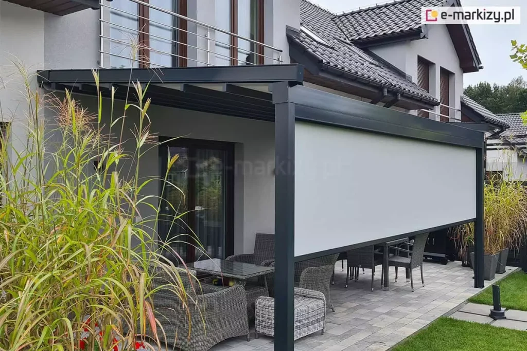 Nowoczesna pergola tarasowa Square o konstrukcji aluminiowej w kolorze antracytowym, wyposażona w system bocznych i frontowych przesłon przeciwsłonecznych typu screen - refleksol Zip L i Zip U. Na zdjęciu widoczna jest białe, materiałowa roleta frontowa, która jest opuszczona do około dwóch trzecich wysokości konstrukcji, osłaniając wnętrze tarasu przed nadmiernym nasłonecznieniem, jednocześnie pozostawiając otwartą przestrzeń przy podłodze. Zadaszenie pergoli jest lekko nachylone i składa się z jasnego poszycia wspartego na ciemnych profilach poprzecznych. Pod pergolą znajduje się zestaw mebli z technorattanu w kolorze szaro-brązowym, a w dalszej części wykonane z tego samego materiału brązowe krzesła. Obok powierzchni tarasu z płyt betonowych widoczny jest zadbany trawnik a po lewej stronie duża donica z ozdobnymi trawami.