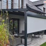 Nowoczesna pergola tarasowa Square o konstrukcji aluminiowej w kolorze antracytowym, wyposażona w system bocznych i frontowych przesłon przeciwsłonecznych typu screen - refleksol Zip L i Zip U. Na zdjęciu widoczna jest białe, materiałowa roleta frontowa, która jest opuszczona do około dwóch trzecich wysokości konstrukcji, osłaniając wnętrze tarasu przed nadmiernym nasłonecznieniem, jednocześnie pozostawiając otwartą przestrzeń przy podłodze. Zadaszenie pergoli jest lekko nachylone i składa się z jasnego poszycia wspartego na ciemnych profilach poprzecznych. Pod pergolą znajduje się zestaw mebli z technorattanu w kolorze szaro-brązowym, a w dalszej części wykonane z tego samego materiału brązowe krzesła. Obok powierzchni tarasu z płyt betonowych widoczny jest zadbany trawnik a po lewej stronie duża donica z ozdobnymi trawami.