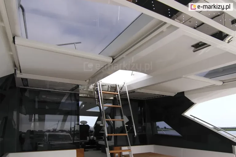 Luksusowe, przeszklone wnętrze górnego pokładu jachtu z zaawansowanym systemem osłonowym. Pod szklanym dachem zamontowano poziome rolety przeciwsłoneczne (system Veranda) w kolorze białym, które poruszają się po aluminiowych prowadnicach. Jedna z rolet jest częściowo zwinięta, odsłaniając widok na niebo. Centralnym punktem kadru są strome schody (drabinka) z chromowanymi poręczami i drewnianymi stopniami, prowadzące do otwieranego luku dachowego. Konstrukcja wnętrza jest biało-czarna, z błyszczącymi panelami i zintegrowanym oświetleniem punktowym LED. W tle widoczne są fotele kapitańskie, ciemne szyby boczne oraz fragment linii brzegowej i wody. Zdjęcie prezentuje estetyczny montaż rolet w niestandardowych, prestiżowych przestrzeniach.