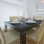 Zdjęcie przedstawia nowoczesną i elegancką jadalnię, w której kluczowym elementem dekoracyjnym i funkcjonalnym są białe żaluzje pionowe, czyli verticale. Przesłaniają one duże okno narożne, tworząc tło z pionowych, gęsto ułożonych pasów materiału, które całkowicie odcinają widok na zewnątrz, jednocześnie rozjaśniając wnętrze. W centralnej części pomieszczenia stoi masywny, prostokątny stół wykonany z ciemnego drewna o wyraźnej teksturze, wokół którego ustawiono sześć nowoczesnych krzeseł z wysokimi oparciami, tapicerowanych gładkim materiałem w kolorze ecru. Na stole znajduje się kompletna zastawa dla czterech osób, składająca się z płaskich białych talerzy, głębszych miseczek oraz polerowanych sztućców. Dekorację stołu dopełniają wysokie, smukłe kieliszki do wina wykonane z dymionego szkła, białe długie świece w srebrnych świecznikach oraz szklana waza z ozdobnymi gałązkami. Na białej ścianie po prawej stronie powieszono duże, nowoczesne lustro o geometrycznym kształcie, składające się z kilku mniejszych tafli, w którym odbija się fragment sufitu i żaluzji. Podłoga w pomieszczeniu wyłożona jest jasnymi panelami o naturalnym odcieniu drewna, co ociepla sterylny, biało-czarny charakter aranżacji.