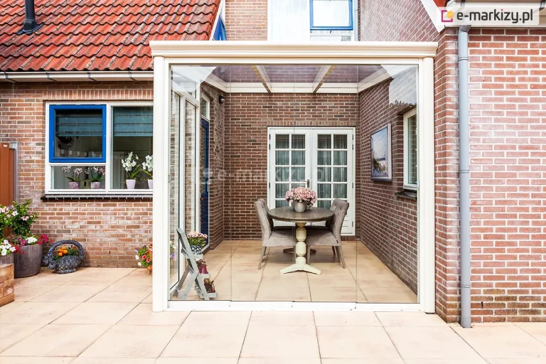 Parterowy budynek z czerwonej cegły z wyjściem na taras z kuchni i salonu. W zagłębieniu pomiędzy ścianami zamocowano zadaszenie tarasu ze szkła z dodatkowymi ścianami bocznymi, systemem szyb przesuwnych bezramowych. Pod zadaszeniem stworzono przytulny kącik jadalniany z owalnym drewnianym stołem, na którym ustawiono wazon z kwiatami oraz czterema wygodnymi krzesłami tapicerowanymi wokół stołu. Obok zadaszenia na tarasie widoczne donice z kwiatami, które tworzą sielski nastrój.