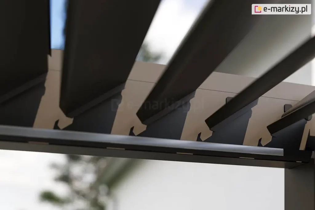 Zbliżenie na mechanizm obrotowy ruchomych lameli sufitowych pergoli bioklimatycznej Rubio w kolorze antracytowym. Zdjęcie wykonane pod kątem od dołu prezentuje sposób osadzenia aluminiowych lameli w bocznej belce ramy. Widoczne są precyzyjne wycięcia w profilu belki, w których zamontowano czarne uchwyty z tworzywa sztucznego, umożliwiające obrót lameli wokół własnej osi. Lamele są ustawione w pozycji półotwartej pod kątem około 45 stopni, co ukazuje ich aerodynamiczny kształt oraz szczeliny pozwalające na regulację nasłonecznienia. Na wewnętrznej krawędzi ramy, poniżej punktów obrotu, zamontowano liniowy profil oświetleniowy LED z mlecznym kloszem. W rozmytym tle widoczne są korony drzew liściastych oraz jasne, dzienne niebo, co podkreśla integrację systemu z naturalnym otoczeniem ogrodu.