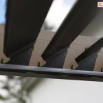 Zbliżenie na mechanizm obrotowy ruchomych lameli sufitowych pergoli bioklimatycznej Rubio w kolorze antracytowym. Zdjęcie wykonane pod kątem od dołu prezentuje sposób osadzenia aluminiowych lameli w bocznej belce ramy. Widoczne są precyzyjne wycięcia w profilu belki, w których zamontowano czarne uchwyty z tworzywa sztucznego, umożliwiające obrót lameli wokół własnej osi. Lamele są ustawione w pozycji półotwartej pod kątem około 45 stopni, co ukazuje ich aerodynamiczny kształt oraz szczeliny pozwalające na regulację nasłonecznienia. Na wewnętrznej krawędzi ramy, poniżej punktów obrotu, zamontowano liniowy profil oświetleniowy LED z mlecznym kloszem. W rozmytym tle widoczne są korony drzew liściastych oraz jasne, dzienne niebo, co podkreśla integrację systemu z naturalnym otoczeniem ogrodu.