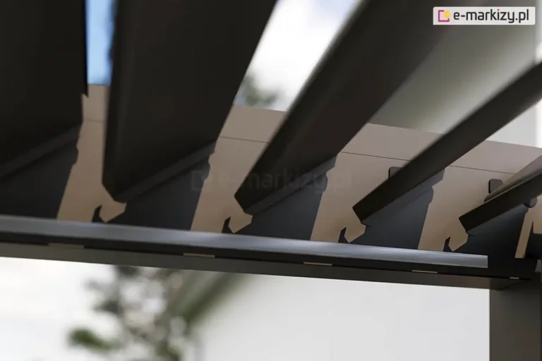 Zbliżenie na mechanizm obrotowy ruchomych lameli sufitowych pergoli bioklimatycznej Rubio w kolorze antracytowym. Zdjęcie wykonane pod kątem od dołu prezentuje sposób osadzenia aluminiowych lameli w bocznej belce ramy. Widoczne są precyzyjne wycięcia w profilu belki, w których zamontowano czarne uchwyty z tworzywa sztucznego, umożliwiające obrót lameli wokół własnej osi. Lamele są ustawione w pozycji półotwartej pod kątem około 45 stopni, co ukazuje ich aerodynamiczny kształt oraz szczeliny pozwalające na regulację nasłonecznienia. Na wewnętrznej krawędzi ramy, poniżej punktów obrotu, zamontowano liniowy profil oświetleniowy LED z mlecznym kloszem. W rozmytym tle widoczne są korony drzew liściastych oraz jasne, dzienne niebo, co podkreśla integrację systemu z naturalnym otoczeniem ogrodu.