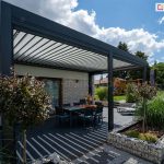 Przydomowy taras pod pergolą z miejscem na opalanie i jedzenie w otoczeniu zieleni i kwiatów