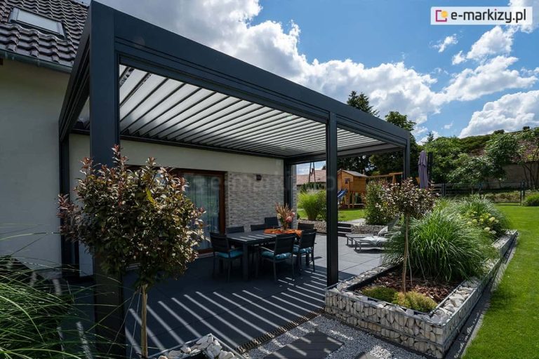 Przydomowy taras pod pergolą z miejscem na opalanie i jedzenie w otoczeniu zieleni i kwiatów. Sielskiego charakteru otoczeniu dodaje zadbany ogród oraz błękitne, letnie niebo z białymi chmurkami.