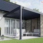 Zdjęcie prezentuje nowoczesną pergolę tarasową Rubio Soft o konstrukcji z aluminium w głębokim odcieniu antracytu, zamontowaną bezpośrednio do białej elewacji domu jednorodzinnego z dachem krytym ciemną dachówką. Zadaszenie pergoli wykonane jest z systemu ruchomych lameli, które na fotografii są częściowo otwarte, co rzuca charakterystyczne, pasiaste cienie na ścianę budynku oraz przeszklone drzwi tarasowe. Konstrukcja posiada jedną boczną ściankę wypełnioną stałymi, poziomymi panelami żaluzjowymi z listewkami w kolorze naturalnego drewna, które zapewniają prywatność i ochronę przed słońcem. Wewnątrz pergoli, na jasnym podłożu, ustawiono biały komplet mebli ogrodowych składający się z fotela, dwuosobowej sofy z szarymi poduszkami oraz minimalistycznego stolika kawowego. Przy przednim słupie nośnym zamontowana jest czarna korba służąca do manualnej regulacji kąta nachylenia lameli dachowych. Na zewnątrz, na betonowym chodniku sąsiadującym z równo przyciętym trawnikiem, stoją dekoracyjne latarnie ogrodowe oraz wysoka, czarna donica z zieloną rośliną o kulistym kształcie. W tle po prawej stronie widoczne są gęste krzewy z fioletowymi kwiatami, co dopełnia estetykę nowoczesnej strefy relaksu. Cała scena jest oświetlona jasnym, dziennym światłem pod lekko zachmurzonym niebem.