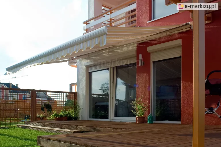 Zdjęcie prezentuje markizę elektryczną Palladio w pełnym rozłożeniu, zamontowaną na elewacji budynku o intensywnym, ceglastym kolorze. Poszycie markizy wykonane jest z tkaniny w delikatne, pionowe pasy w odcieniach bieli, jasnej żółci oraz szarości, wykończone klasyczną, pofalowaną falbaną o identycznym wzorze. Biała konstrukcja kasety oraz profilu czołowego harmonizuje z białymi ramami dużych drzwi tarasowych, w których odbija się fragment sąsiedniej zabudowy. Nad markizą widoczny jest balkon z prostą, nowoczesną balustradą łączącą białe panele z drewnianymi pochwytami. Taras wykonany z ciemnobrązowych desek drewnianych przylega bezpośrednio do wypielęgnowanego trawnika. Po prawej stronie kadru znajduje się ażurowa, drewniana konstrukcja pergoli, przy której ustawiono dziecięcy wózek spacerowy. Aranżację uzupełniają donice z roślinami oraz zielona konewka stojąca przy wejściu. Perspektywa zdjęcia ujęta z boku podkreśla głębokość wysięgu markizy oraz jej funkcjonalność w tworzeniu strefy cienia nad miejscem wypoczynku.