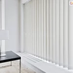 Jasne i przestronne wnętrze biurowe, w którym zainstalowano żaluzje pionowe osłaniające duże przeszklenia. Materiałowe lamele w kolorze czystej bieli zwisają pionowo od sufitu aż do poziomu szerokiego parapetu, tworząc gęstą i uporządkowaną strukturę przesłony. Żaluzje pionowe są ustawione w pozycji zamkniętej, z delikatnie nachylonymi pasami materiału, co pozwala na skuteczne rozproszenie światła dziennego docierającego do środka. Po lewej stronie kadru znajduje się mniejsze okno, również wyposażone w ten sam system osłonowy. Na pierwszym planie, po lewej stronie, stoi czarne biurko o matowym blacie z nowoczesną lampką biurkową, która posiada prostokątny, biały abażur i metalową podstawę. Podłoga wykonana jest z jasnych paneli o wyraźnym rysunku drewna. Cała kompozycja utrzymana jest w minimalistycznym, niemal całkowicie białym stylu, a gra światła i cienia na lamelach podkreśla trójwymiarowość żaluzji.