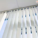 Zbliżenie od dołu na żaluzje pionowe zamontowane pod sufitem wzdłuż szerokiego przeszklenia. Pionowe lamele o szerokości 12,7 cm wykonane są z gładkiego materiału w odcieniu złamanej bieli. Każdy pas tkaniny jest zamocowany do górnej szyny za pomocą białych uchwytów, które umożliwiają ich obracanie wokół własnej osi. Na zdjęciu żaluzje pionowe są częściowo obrócone pod kątem, co tworzy rytmiczny wzór z powtarzających się geometrycznych płaszczyzn materiału oraz przerw między nimi. Przez szczeliny wpada rozproszone światło dzienne, ukazując fragmenty ramy okiennej oraz błękitne niebo na zewnątrz. Konstrukcja szyny jest biała i przylega bezpośrednio do jasnego sufitu kasetonowego z widocznymi liniami podziału. Perspektywa żabia podkreśla wysokość systemu osłonowego oraz precyzyjne ułożenie poszczególnych elementów tkaniny, które tworzą pionowe pasy światła i cienia w pomieszczeniu.