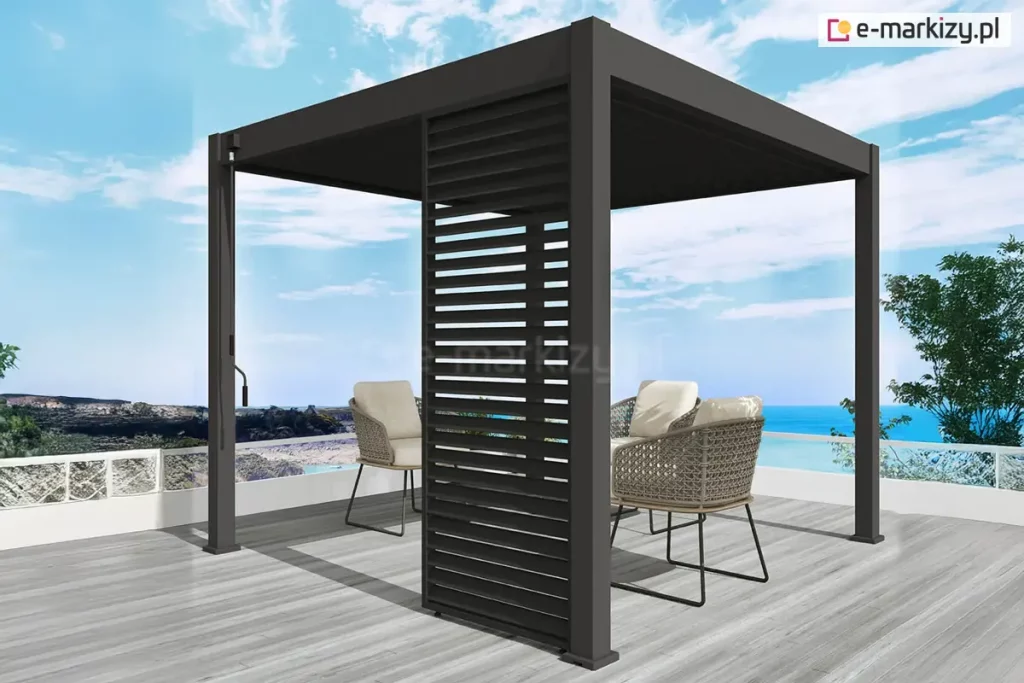 Antracytowa wolnostojąca pergola lamelowa Marlo o kwadratowej podstawie, zainstalowana na tarasie z widokiem na morze. Konstrukcja zadaszenia posiada dach z ruchomych lamel ustawionych w pozycji całkowicie zamkniętej oraz pionowy panel żaluzjowy zamontowany w jednym z narożników, który zapewnia osłonę przed bocznym wiatrem i słońcem. Przy jednym ze słupów nośnych widoczna jest metalowa korba służąca do ręcznego sterowania mechanizmem dachu. Pod pergolą, na podłożu z jasnoszarych desek tarasowych, ustawiono dwa nowoczesne fotele z ażurowym, plecionym oparciem i jasnymi poduszkami. Taras ograniczony jest białą barierką z przeszkleniami, za którą rozciąga się nadmorski krajobraz z linią brzegową oraz jasnobłękitne niebo z delikatnymi pasmami chmur.