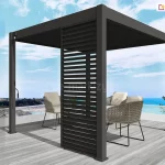 Antracytowa wolnostojąca pergola lamelowa Marlo o kwadratowej podstawie, zainstalowana na tarasie z widokiem na morze. Konstrukcja zadaszenia posiada dach z ruchomych lamel ustawionych w pozycji całkowicie zamkniętej oraz pionowy panel żaluzjowy zamontowany w jednym z narożników, który zapewnia osłonę przed bocznym wiatrem i słońcem. Przy jednym ze słupów nośnych widoczna jest metalowa korba służąca do ręcznego sterowania mechanizmem dachu. Pod pergolą, na podłożu z jasnoszarych desek tarasowych, ustawiono dwa nowoczesne fotele z ażurowym, plecionym oparciem i jasnymi poduszkami. Taras ograniczony jest białą barierką z przeszkleniami, za którą rozciąga się nadmorski krajobraz z linią brzegową oraz jasnobłękitne niebo z delikatnymi pasmami chmur.