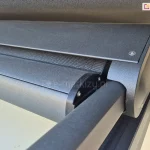 Szczegółowe zbliżenie na aluminiową kasetę markizy dachowej Veranda w kolorze antracytowym z matowym wykończeniem. Zdjęcie prezentuje owalna kasetę, z której wysuwa się ciemna tkanina osłonowa z belką przednią. Obok nich widoczna jest prowadnica boczna, w której przesuwa się belka dolna napinając tkaninę. Konstrukcja charakteryzuje się nowoczesnym, minimalistycznym designem z wyraźnie zaznaczonymi łączeniami elementów i śrubami montażowymi. Kaseta jest zamontowana samonośnie, na prowadnicach, tuż pod krawędzią dachu lub nadproża, a w dolnej części kadru widoczny jest fragment czarnego profilu konstrukcji zadaszenia. Precyzyjne spasowanie elementów zapewnia szczelność i ochronę mechanizmu przed czynnikami atmosferycznymi.