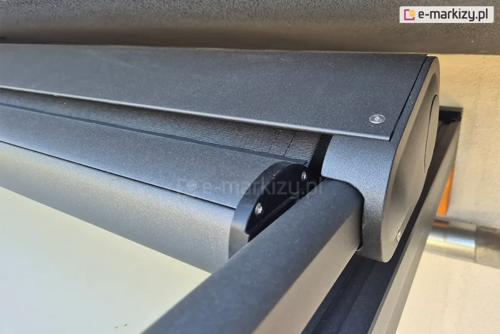 Szczegółowe zbliżenie na aluminiową kasetę markizy dachowej w kolorze antracytowym z matowym wykończeniem. Zdjęcie prezentuje owalna kasetę, z której wysuwa się ciemna tkanina osłonowa z belką przednią. Obok nich widoczna jest prowadnica boczna, w której przesuwa się belka dolna napinając tkaninę. Konstrukcja charakteryzuje się nowoczesnym, minimalistycznym designem z wyraźnie zaznaczonymi łączeniami elementów i śrubami montażowymi. Kaseta jest zamontowana samonośnie, na prowadnicach, tuż pod krawędzią dachu lub nadproża, a w dolnej części kadru widoczny jest fragment czarnego profilu konstrukcji zadaszenia. Precyzyjne spasowanie elementów zapewnia szczelność i ochronę mechanizmu przed czynnikami atmosferycznymi.