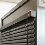 Zbliżenie na górny profil oraz mechanizm montażowy żaluzji poziomej C50 Slim o nowoczesnym designie. Zdjęcie prezentuje czarne, aluminiowe lamele o wygiętym profilu w kształcie litery C, zawieszone na precyzyjnym systemie drabinek sznurkowych. Górna rynna żaluzji jest osłonięta elegancką, szeroką maskownicą w kolorze antracytowym o matowym wykończeniu, która jest stabilnie zamontowana do ramy okiennej. Na bocznej krawędzi maskownicy widoczny jest subtelny, czerwony logotyp producenta oraz metalowe elementy wykończeniowe. Detal podkreśla wysoką jakość spasowania komponentów oraz techniczny charakter systemu osłonowego, idealnie komponującego się z nowoczesną stolarką otworową. Tło stanowi neutralna, jasna ściana oraz fragment profilu aluminiowego, co pozwala w pełni skupić uwagę na konstrukcji samej żaluzji.