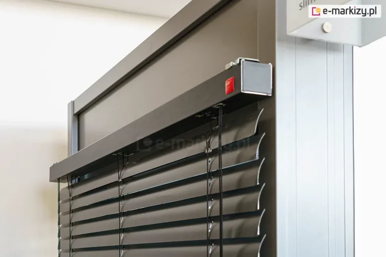 Zbliżenie na górny profil oraz mechanizm montażowy żaluzji poziomej C50 Slim o nowoczesnym designie. Zdjęcie prezentuje czarne, aluminiowe lamele o wygiętym profilu w kształcie litery C, zawieszone na precyzyjnym systemie drabinek sznurkowych. Górna rynna żaluzji jest osłonięta elegancką, szeroką maskownicą w kolorze antracytowym o matowym wykończeniu, która jest stabilnie zamontowana do ramy okiennej. Na bocznej krawędzi maskownicy widoczny jest subtelny, czerwony logotyp producenta oraz metalowe elementy wykończeniowe. Detal podkreśla wysoką jakość spasowania komponentów oraz techniczny charakter systemu osłonowego, idealnie komponującego się z nowoczesną stolarką otworową. Tło stanowi neutralna, jasna ściana oraz fragment profilu aluminiowego, co pozwala w pełni skupić uwagę na konstrukcji samej żaluzji.