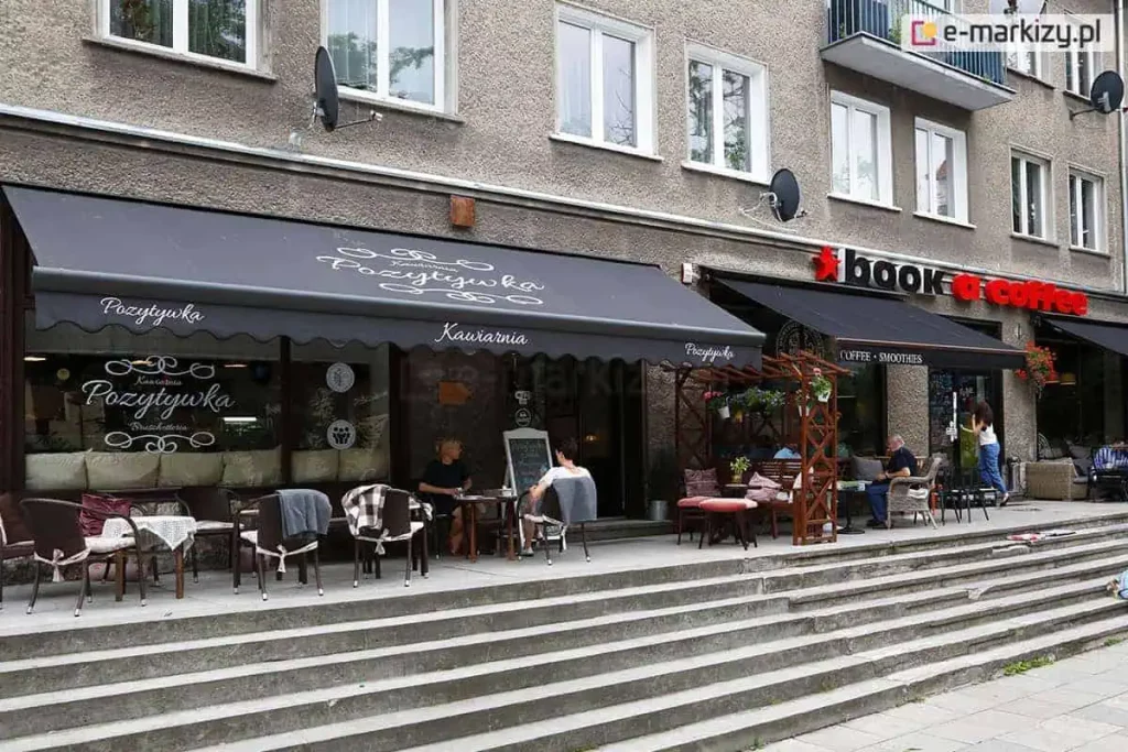 Czarna markiza reklamowa z ozdobnymi białymi napisami, osłaniająca ogródek kawiarniany w parterze miejskiej kamienicy. Na głównym poszyciu oraz na pofalowanej falbanie widnieje estetyczny nadruk logo kawiarni "Pozytywka" wykonany ozdobną czcionką z dekoracyjnymi zawijasami. Obok znajduje się kolejna, mniejsza osłona pod szyldem "book a coffee". Pod markizami ustawiono liczne stoliki, wiklinowe i metalowe krzesła z poduszkami oraz koce dla gości. Witryny lokalu są przeszklone, a na jednej z nich powtórzono logotyp kawiarni. Przed wejściem do lokali znajdują się szerokie, betonowe schody prowadzące na poziom chodnika. W ogródku widoczni są klienci siedzący przy stolikach oraz drewniane konstrukcje pergoli z donicami kwiatowymi. Elewacja budynku jest szara, o chropowatej strukturze, z widocznymi powyżej oknami mieszkalnymi i antenami satelitarnymi. Całość tworzy przytulną przestrzeń komercyjną zintegrowaną z miejską architekturą.