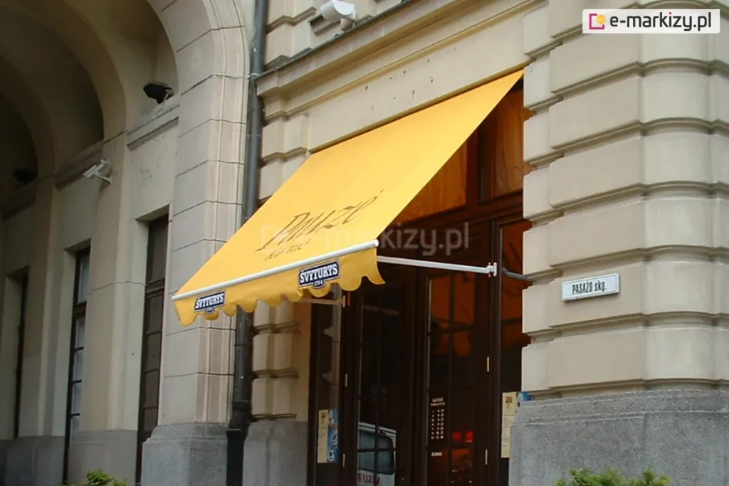 Kawiarnia usytuowana w zabytkowym budynku w Wilnie, której wejście zadaszone zostało markizą sklepową Italia Aluprof z białymi ramionami mocowanymi do ramy drzwi wejściowych oraz żółtą tkaniną poszycia z dużym nadrukiem oraz reklamą na wyciętej w półkola falbanie.