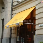 Kawiarnia usytuowana w zabytkowym budynku w Wilnie, której wejście zadaszone zostało markizą sklepową Italia Aluprof z białymi ramionami mocowanymi do ramy drzwi wejściowych oraz żółtą tkaniną poszycia z dużym nadrukiem oraz reklamą na wyciętej w półkola falbanie.