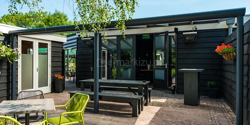 Zdjęcie prezentuje szerokie zadaszenie tarasowe Nebbiolo marki Deponti, zamontowane wzdłuż elewacji parterowego budynku o ciemnej, czarnej fasadzie wykonanej z poziomych paneli. Konstrukcja opiera się na antracytowym, aluminiowym stelażu z długą belką frontową, wspartą na pionowych słupach. Pod zadaszeniem zaaranżowano przestrzeń kawiarniano-restauracyjną z czarnym, drewnianym stołem i ławami oraz mniejszymi stolikami z nowoczesnymi, ażurowymi krzesłami w kolorze jaskrawej limonki. Taras wyłożony jest postarzaną kostką brukową w odcieniach brązu. Na ścianie budynku widoczne są zamontowane promienniki ciepła oraz zawieszone donice z kwitnącymi czerwonymi kwiatami. W tle, przez otwarte przeszklone drzwi, widać wnętrze lokalu, a nad całością konstrukcji pochylają się gałęzie brzozy z zielonymi liśćmi na tle jasnego nieba.