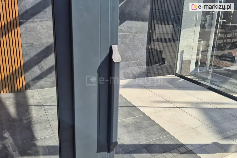 Zbliżenie klamki zewnętrznej systemu szyb przesuwnych ramowego Sotro. W tle widoczny taras pokryty różnego rodzaju płytami ozdobnymi. Na ścianie płyty grafitowej i panel z drewnianymi lamelami po lewej.