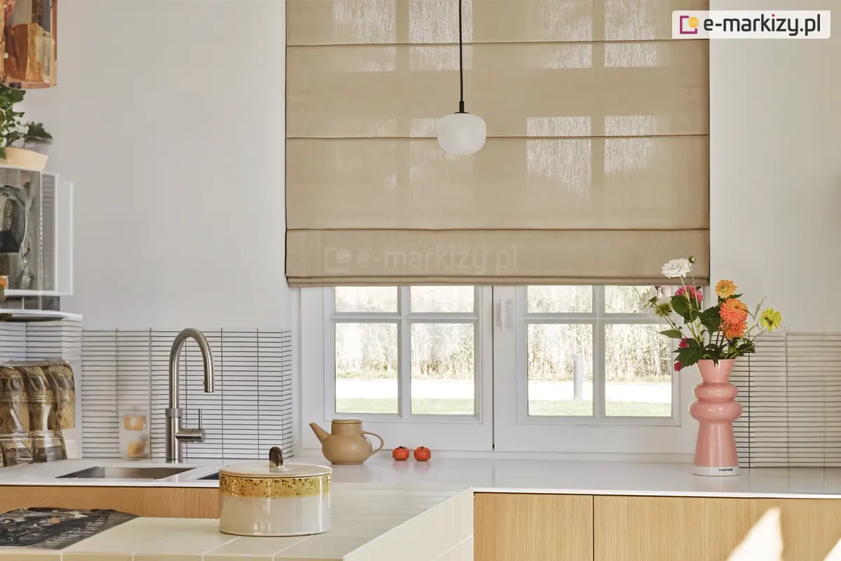 Zdjęcie prezentuje jasną i nowoczesną kuchnię, w której głównym elementem dekoracyjnym okna jest beżowa roleta rzymska. Tkanina rolety ma delikatny, pionowy splot i jest półtransparentna, co pozwala na przenikanie miękkiego światła dziennego do wnętrza, jednocześnie zapewniając prywatność. Roleta jest częściowo opuszczona, a jej struktura jest wyraźnie podzielona poziomymi fiszbinami na kilka równych sekcji, przy czym dolna część układa się w estetyczną, płaską zakładkę. Okno pod roletą jest białe, dwuskrzydłowe, z klasycznymi szprosami dzielącymi szyby na mniejsze kwadraty. Przez przeszklenia widać fragment ogrodu lub zarośli w jasnych, naturalnych barwach. Blat kuchenny wykonany jest z gładkiego, białego materiału, na którym ustawiono dekoracyjne i funkcjonalne przedmioty. Po prawej stronie znajduje się wysoki, różowy wazon o geometrycznym, bąbelkowym kształcie, w którym umieszczono bukiet kolorowych kwiatów, w tym pomarańczowe i różowe cynie oraz białe margerytki. Obok wazonu, na ścianie, widać fragment białych płytek ułożonych w wąskie, poziome paski. W centralnej części blatu, pod oknem, stoi beżowy, ceramiczny imbryk oraz dwa małe, czerwone owoce przypominające pomidory koktajlowe. Po lewej stronie widoczny jest zlewozmywak z nowoczesną, srebrną baterią o wysokiej wylewce. Na pierwszym planie widać fragment kuchennej wyspy wyłożonej jasnymi płytkami, na której stoi ceramiczny pojemnik z ozdobną, nakrapianą pokrywką. Szafki kuchenne mają fronty w kolorze jasnego drewna, co w połączeniu z bielą ścian i beżem rolety tworzy spójną i ciepłą aranżację. Z sufitu, na wysokości górnej krawędzi okna, zwisa minimalistyczna lampa z okrągłym, mlecznobiałym kloszem.