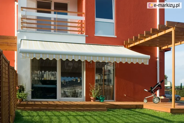 Zdjęcie prezentuje nowoczesny budynek jednorodzinny o intensywnie pomarańczowej elewacji, na którym zainstalowano markizę tarasową Palladio w kasecie. Poszycie markizy wykonane jest z tkaniny w delikatne, pionowe pasy w odcieniach bieli, jasnej żółci i szarości, zakończone klasyczną, pofalowaną falbaną. Markiza jest w pełni rozłożona, osłaniając duże przeszklenia tarasowe w białych ramach, w których odbija się fragment sąsiedniej zabudowy. Konstrukcja markizy jest polakierowana na biało, co harmonizuje z balustradą balkonu znajdującego się na piętrze oraz stolarką okienną. Taras wykonany z naturalnych desek drewnianych jest zintegrowany z niewielkim trawnikiem. Po prawej stronie widoczna jest ażurowa, drewniana pergola wsparta na metalowym słupie, a pod nią dziecięcy wózek spacerowy. Aranżację uzupełniają rośliny w donicach oraz zielona konewka ustawiona przy progu drzwi przesuwnych. Całość kompozycji, ujęta w pełnym słońcu, podkreśla funkcjonalność osłony przeciwsłonecznej w codziennym użytkowaniu przydomowej strefy relaksu.