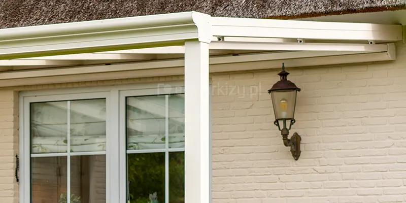 Zbliżenie na detal wykończeniowy rynny przedniej w zadaszeniu tarasowym model Terano. Konstrukcja wykonana jest z profili aluminiowych lakierowanych na kolor kremowy. Widoczny jest profil rynnowy o klasycznym, dekoracyjnym kształcie z delikatnymi żłobieniami, który łączy się z pionowym słupem nośnym o kwadratowym przekroju. Miejsce styku rynny ze słupem zostało zabezpieczone dopasowaną maskownicą narożną, przytwierdzoną za pomocą widocznych wkrętów w kolorze profilu. Zadaszenie jest przymocowane do jasnej ściany budynku z cegły, na której zawieszona jest klasyczna latarnia ogrodowa w stylu retro. Ponad konstrukcją widoczny jest fragment krawędzi dachu krytego strzechą oraz biała rama okienna z podziałami szprosowymi. Wypełnienie dachu zadaszenia stanowią przezroczyste panele,.