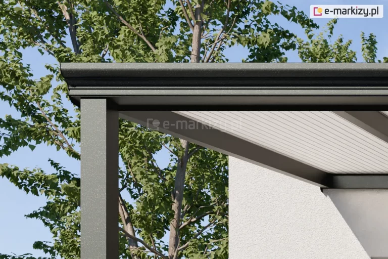 Zbliżenie na górny narożnik nowoczesnego zadaszenia tarasowego Lunaro, skupiające się na detalu klasycznej rynny przedniej oraz sposobie jej łączenia ze słupem nośnym. Konstrukcja wykonana jest z aluminium w kolorze ciemnego antracytu o drobnoziarnistej teksturze. Frontowa rynna posiada eleganckie, poziome przetłoczenia i lekko zaokrąglony profil. Pod rynną widoczny jest fragment dachu z wypełnieniem z białego, poliwęglanu komorowego, który jest osadzony w aluminiowych profilach. W tle znajduje się jasna elewacja budynku oraz gęste korony zielonych drzew liściastych na tle czystego, błękitnego nieba.