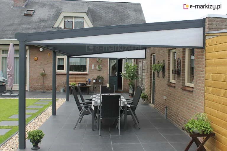 Zadaszenie Bosco Deponti Lunaro z poliwęglanem z dodatkowymi klinami na obu bokach konstrukcji osłania ustawiony na tarasie z dużych grafitowych płyt stół jadalny z ośmioma antracytowymi krzesłami. W tle widoczne ceglane ściany, zielone drzwi wejściowe do domu z dużą szybą, lekko uchylone oraz rośliny doniczkowe poustawiane w różnych miejscach. Po lewej w głębi widoczny zwinięty duży parasol ogrodowy z pokrowcem.