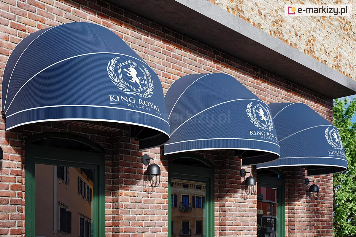 Trzy markizy koszowe półokrągłe z niebieskim poszyciem, prostą falbaną oraz białym wykończeniem dopasowanym kolorystycznie do nadrukowanego na markizach dużego logo hotelu. Markizy umieszczone symetrycznie na ścianie z czerwonej cegły z nowoczesnymi lampami pomiędzy markizami, które osłaniają trzy okna o zielonej ramie.