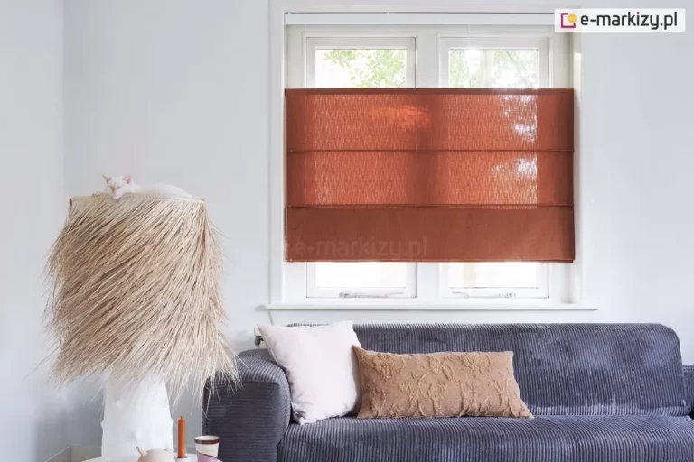 Zdjęcie przedstawia jasne, nowocześnie urządzone wnętrze salonu, w którym centralnym punktem jest okno z zamontowaną roletą rzymską w kolorze terakotowym lub rdzawym brązie. Roleta wykonana jest z półtransparentnej tkaniny o wyraźnym, pionowym splotu, który delikatnie przepuszcza naturalne światło dzienne, tworząc przytulną atmosferę. Osłona jest podciągnięta do połowy wysokości białej ramy okiennej, a jej materiał jest podzielony poziomymi fiszbinami na trzy równe sekcje, co nadaje jej strukturalny wygląd. Okno jest dwuskrzydłowe, z widocznymi klamkami i białym parapetem. Poniżej okna znajduje się fragment ciemnoszarej, tapicerowanej sofy o sztruksowej fakturze, na której ułożone są dwie poduszki: jedna gładka, jasnokremowa, a druga podłużna w kolorze brązowym z wypukłym, ozdobnym wzorem. Po lewej stronie kadru stoi wysoka, dekoracyjna lampa podłogowa z unikalnym abażurem wykonanym z gęstych, jasnych włókien przypominających trawę lub słomę. Na szczycie abażuru leży biały kot, zwinięty w kłębek. Ściany pomieszczenia są gładkie i białe, co kontrastuje z intensywnym kolorem rolety i ciemną sofą. Światło wpadające przez okno oświetla fakturę materiałów, podkreślając detale tkanin i naturalny charakter aranżacji.