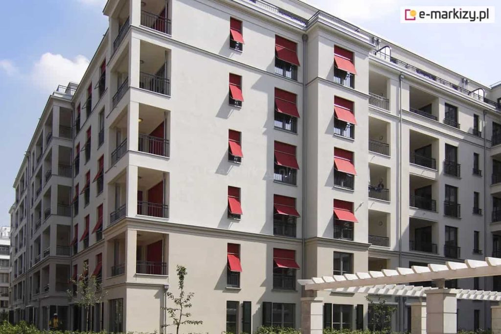 Zdjęcie prezentuje fasadę nowoczesnego, wielorodzinnego budynku mieszkalnego o jasnej, beżowo-szarej elewacji, na której zainstalowano liczne markizolety w intensywnym czerwonym kolorze. Osłony przeciwsłoneczne są zamontowane w pionowych rzędach okien i znajdują się w różnym stopniu rozwinięcia, przy czym większość z nich ma wysuniętą dolną część na ruchomych ramionach, tworząc charakterystyczny pochyły daszek. Konstrukcja markizolet łączy cechy pionowej rolety materiałowej z funkcją markizy, co pozwala na skuteczne zacienienie wnęk okiennych przy zachowaniu dopływu naturalnego światła i powietrza do wnętrz. Budynek posiada liczne loggie oraz balkony z ciemnymi, ażurowymi balustradami, a na pierwszym planie widoczna jest biała pergola ogrodowa oraz fragmenty niskiej zieleni. Intensywna czerwień tkanin technicznych stanowi wyraźny akcent kolorystyczny, kontrastujący z neutralną barwą ścian i błękitem nieba.