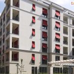 Zdjęcie prezentuje fasadę nowoczesnego, wielorodzinnego budynku mieszkalnego o jasnej, beżowo-szarej elewacji, na której zainstalowano liczne markizolety w intensywnym czerwonym kolorze. Osłony przeciwsłoneczne są zamontowane w pionowych rzędach okien i znajdują się w różnym stopniu rozwinięcia, przy czym większość z nich ma wysuniętą dolną część na ruchomych ramionach, tworząc charakterystyczny pochyły daszek. Konstrukcja markizolet łączy cechy pionowej rolety materiałowej z funkcją markizy, co pozwala na skuteczne zacienienie wnęk okiennych przy zachowaniu dopływu naturalnego światła i powietrza do wnętrz. Budynek posiada liczne loggie oraz balkony z ciemnymi, ażurowymi balustradami, a na pierwszym planie widoczna jest biała pergola ogrodowa oraz fragmenty niskiej zieleni. Intensywna czerwień tkanin technicznych stanowi wyraźny akcent kolorystyczny, kontrastujący z neutralną barwą ścian i błękitem nieba.