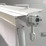 Zbliżenie konstrukcji markizy Italia kasetowa z napędem ręcznym w kolorze białym z białym poszyciem. Widoczna od prawego boku kaseta z lekko wysunięta belką przednią i umocowanym w niej ramieniem. Do boku kasety przymocowana przekładnia korbowa z zaczepem do sterowania ręcznego za pomocą korby. W tle nad kasetą widoczne mocowania do sufitu. Od prawego mocowania oznaczonego okrągłym znacznikiem poprowadzono w prawo zieloną kreskę do umieszczonego na szarym tle napisu "uchwyt sufitowy". Poniżej, od brzegu kasety, na której skraju umieszczono okrągły znacznik poprowadzono w dół i na prawo kreskę do umieszczonego na szarym tle napisu "niewielka kaseta (126x119mm)".