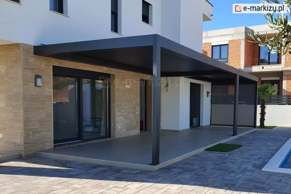 Pergola SB400 ustawiona na tarasie domu z jasną elewacją nad tarasem betonowym w pobliżu basenu oraz podwórka wyłożonego kostką. Jedna ze ścian osłonięta została refleksolem.