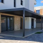 Pergola SB400 ustawiona na tarasie domu z jasną elewacją nad tarasem betonowym w pobliżu basenu oraz podwórka wyłożonego kostką. Jedna ze ścian osłonięta została refleksolem.