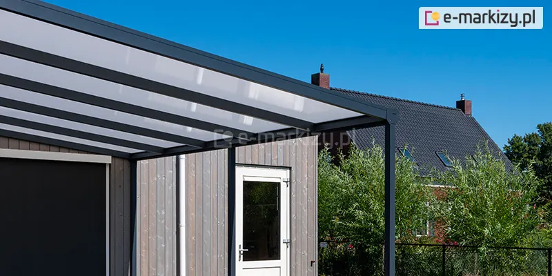 Zdjęcie przedstawia nowoczesne zadaszenie tarasowe o konstrukcji aluminiowej Nebbiolo Deponti w kolorze ciemnoantracytowym. System składa się z wytrzymałych profili nośnych oraz równolegle rozmieszczonych krokwi dachowych, w których osadzono płyty z poliwęglanu komorowego w kolorze mlecznym. Konstrukcja charakteryzuje się prostą, minimalistyczną formą i jest zamontowana przy budynku o elewacji wykonanej z pionowych paneli drewnianych w odcieniu szaro-brązowym, w której znajdują się białe drzwi z przeszkleniem. Po lewej stronie konstrukcji widoczna jest pionowa roleta boczna w ciemnym kolorze, zapewniająca dodatkową osłonę. W tle znajduje się fragment sąsiedniego domu z ciemną dachówką oraz zadbana zieleń ogrodowa i drzewa, a całość ujęcia dopełnia czyste, błękitne niebo w pełnym świetle dziennym.